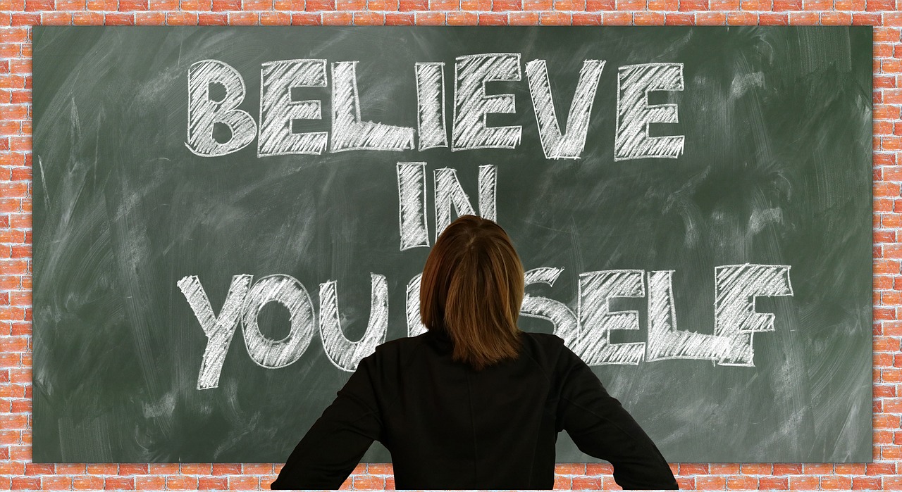 Back of woman's head looking at a green chalk board with the words Believe in Yourself written in bold block letters.